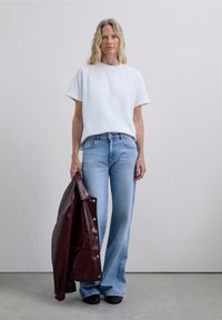 Donna bionda che indossa una maglietta bianca e jeans azzurri chiari tiene una giacca di pelle rossa scura nella mano sinistra, in piedi su un pavimento grigio.