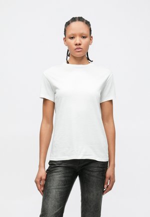 Ung person med flettet hår som har på seg en ensfarget hvit t-skjorte og svarte teksturerte bukser, står foran en hvit bakgrunn.