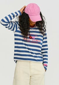 Modro-bílé pruhované tričko s dlouhým rukávem, na kterém je růžový vyšitý text a květiny. V kombinaci se smetanovými kalhotami a růžovou čepicí.