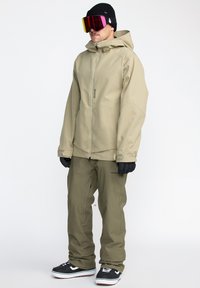 Veste d'extérieur beige avec fermeture zip, associée à un pantalon vert olive. Gants noirs et bonnet, complétés par des lunettes de ski miroir.