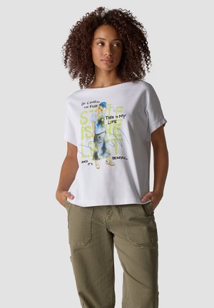 Woman with curly hair wearing a white graphic t-shirt and olive green pants, hands in pockets, looking slightly to the side.