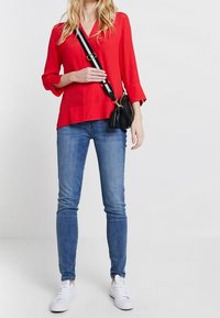 Donna con una blusa rossa, jeans blu, sneakers bianche e una borsa a tracolla nera con una tracolla a righe, in piedi contro uno sfondo neutro.