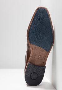 Semelle en cuir marron de chaussure habillée avec des zones de grip en caoutchouc bleu et noir, taille 42, arborant le logo "bugatti" et un logo d'amortisseur.