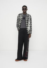 Καρό πουκάμισο σε μαύρο και άσπρο με γυαλιστερές λεπτομέρειες, γκρι T-shirt με σχέδιο αστεριού, μαύρο παντελόνι φαρδιάς γραμμής με κόκκινα και λευκά σχέδια, και μαύρα παπούτσια.