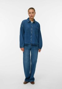 Camicia in denim con colletto, bottoni e tasca sul petto; abbinata a jeans in denim blu a gamba larga. Entrambi presentano un colore blu uniforme.