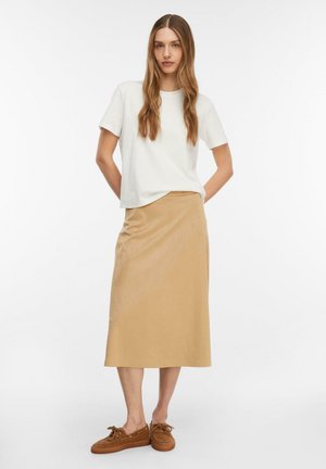 Kobieta stojąca z długimi włosami, ubrana w białą koszulę z krótkim rękawem, beżową spódnicę midi i brązowe sznurowane buty, na tle jednolitego białego tła.