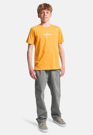 T-shirt en coton jaune avec un logo blanc sur la poitrine, associé à un jean gris. Le modèle porte des chaussures grises avec des semelles blanches.