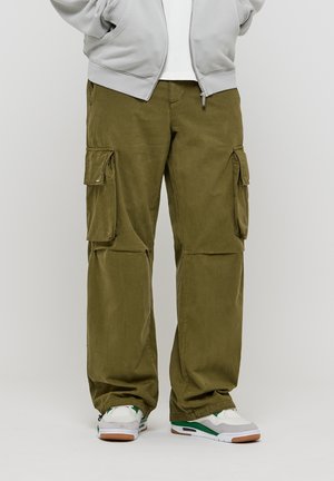 Person trägt olivgrüne Cargohosen mit mehreren Taschen, weiße und grüne Sneakers sowie eine hellgraue Zip-up-Jacke.