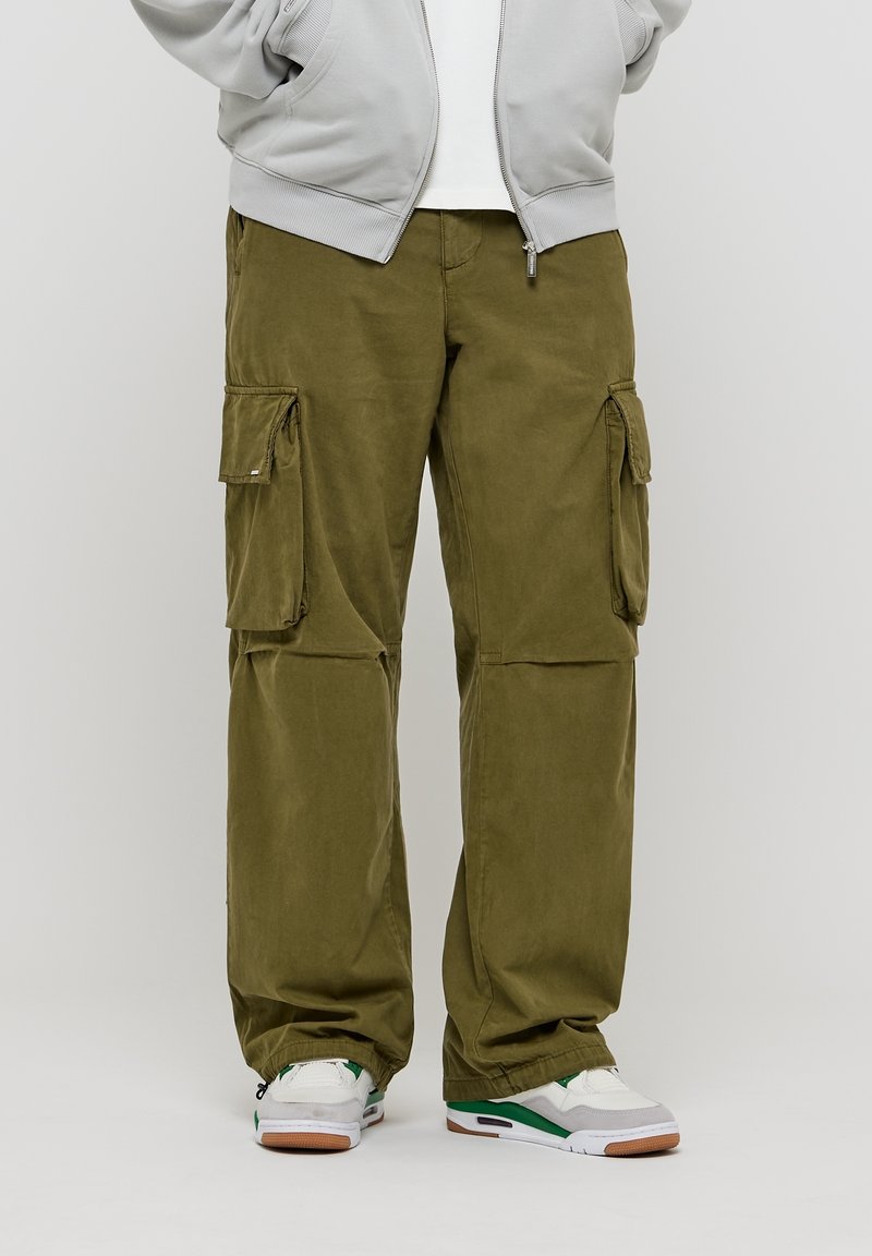 Personne portant un pantalon cargo vert olive avec plusieurs poches, des baskets blanches et vertes, et une veste zippée gris clair.