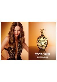 La botella del perfume Roberto Cavalli Sweet Ferocious presenta una forma ovalada dorada con un patrón de rayas de tigre y una tapa decorativa, todo ello sobre un fondo cálido.