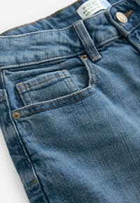 Jeans de mezclilla azul con una cintura de altura media, que presentan un bolsillo frontal y acentos de botón de latón. La tela tiene una textura suave y desgastada.