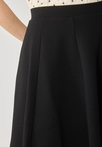 Femme portant une jupe noire texturée taille haute et un chemisier blanc à pois noirs, bras reposant sur la hanche.