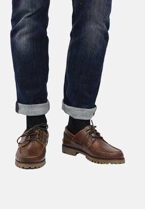 Braune Lederschuhe mit strukturierter Oberfläche, runder Zehenform und kontrastierender Gummisohle; kombiniert mit dunklen Jeans, zeigen die aufgerollten Säume ein graues Futter.