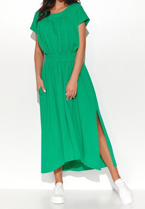 Nainen, jolla on kirkkaanvihreä midi-mekko, lyhyet hihat, joustava vyötärö, sivuhalkio ja valkoiset lenkkarit, yksinkertaisen valkoisen taustan edessä.