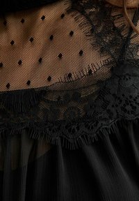 Vestido negro con una superposición de malla de lunares, un escote de encaje intrincado y tela plisada suave. Detalles que incluyen bordes festoneados y texturas finas.