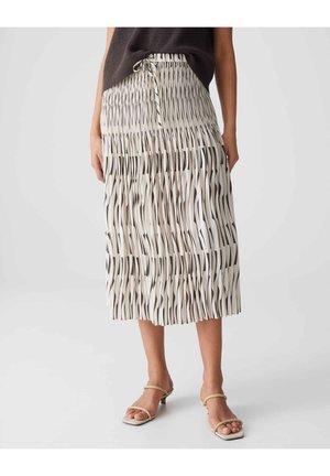 Frau trägt einen Midi-Faltenrock mit schwarz-weißen vertikalen wellenförmigen Streifen und beigen Sandaletten mit Absatz, steht vor einem schlichten Hintergrund.