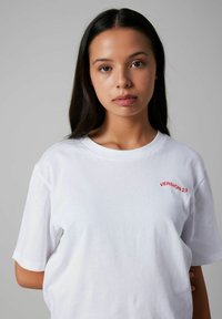 Camiseta de algodón blanca con mangas cortas, que presenta el texto rojo "VERSIÓN 2.0" en el pecho. Ajuste simple y relajado sin patrones adicionales.