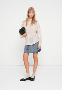 Blusa beige leggera con ricami floreali, abbinata a una gonna mini in denim, calze color crema e ballerine nere, mentre tiene una piccola borsa nera.