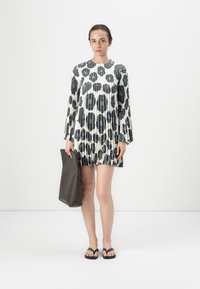 Plissiertes Kleid mit weißem Grund und schwarzem abstraktem Blumenmuster, mit langen Ärmeln. Das Modell hält eine dunkelgraue Tote-Bag. Schwarze Flip-Flops.