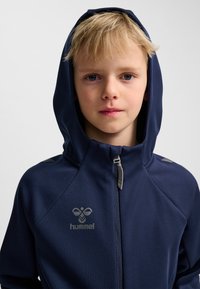 Mörkblå hoodie i slät tyg, med dragkedja framtill, huva och en logotyp. Inkluderar silverdetaljer på axlarna.