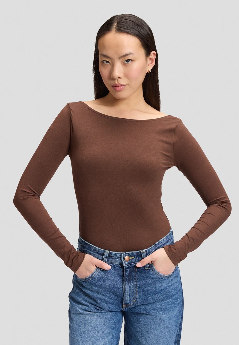 Jeune femme aux longs cheveux foncés porte un haut marron à manches longues et un jean bleu, debout les mains dans les poches devant un fond uni.