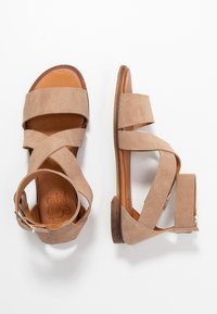 Sandalias de ante beige con tiras entrelazadas, puntera abierta y hebilla en el tobillo. Textura suave, suela plana y diseño minimalista.