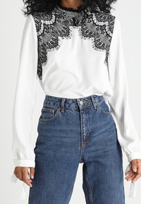 Blouse blanche avec un empiècement en dentelle noire et des manches longues à poignets froncés, associée à un jean bleu taille haute.