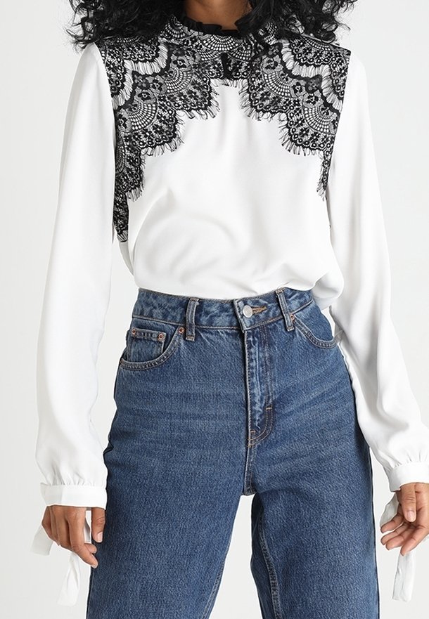 Blouse blanche avec un empiècement en dentelle noire et des manches longues à poignets froncés, associée à un jean bleu taille haute.