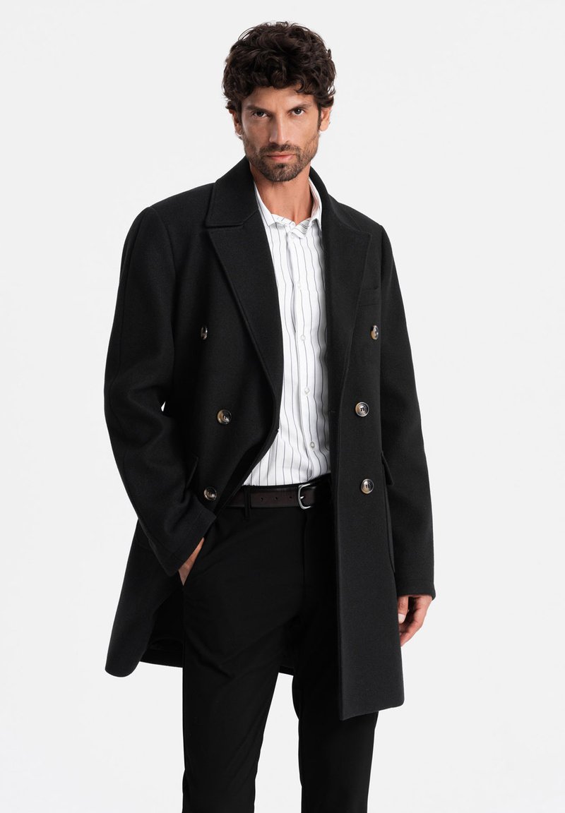 Cappotto doppiopetto in lana nera con bottoni metallici, dotato di colletto a lancia e vestibilità rilassata, indossato sopra una camicia a righe e pantaloni neri.