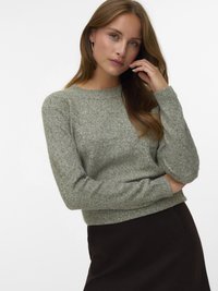 Giovane donna con lunghi capelli castano chiaro che indossa orecchini a cerchio argento, un maglione lavorato a maglia grigio chiaro e una gonna scura, che si tocca il viso.