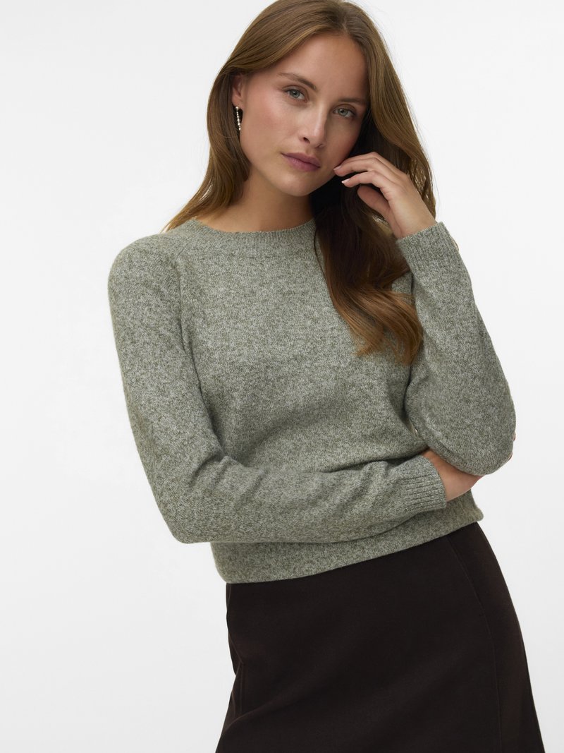 Giovane donna con lunghi capelli castano chiaro che indossa orecchini a cerchio argento, un maglione lavorato a maglia grigio chiaro e una gonna scura, che si tocca il viso.