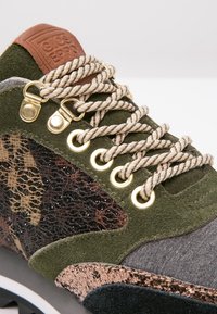 Gros plan d'une basket en daim vert avec des lacets tressés beige, des œillets dorés, un panneau en mesh camouflage et une étiquette en cuir marron sur la languette.