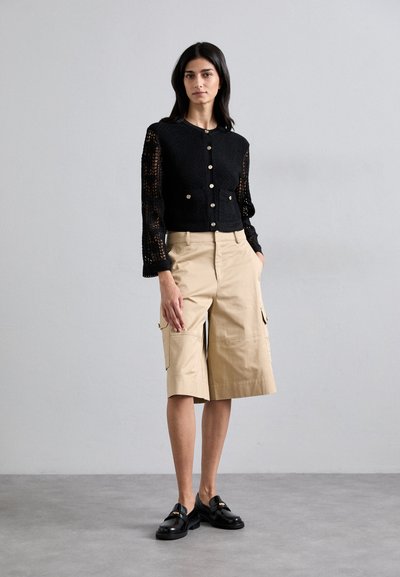 Cardigan noir au crochet avec fermeture à boutons et manches en dentelle, associé à un short cargo beige arrivant au genou et des mocassins noirs. Look moderne et texturé.