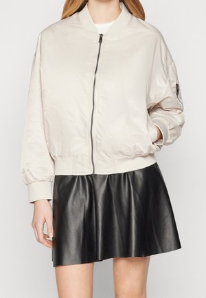 Kobieta w jasnobeżowej bomberce z przednim zamkiem błyskawicznym, kieszenią na rękawie oraz czarnej, skórzanej, rozkloszowanej mini spódnicy.