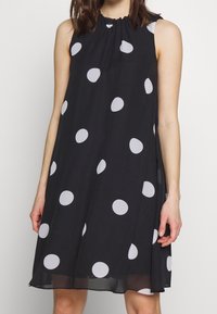 Svart ärmlös klänning med en insnörd halsringning, prydd med stora vita prickar, tillverkad av lätt tyg, knälång design.