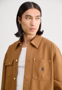 Cappotto marrone con colletto, bottoni frontali e tasche. Presenta dettagli in metallo dorato e un'etichetta con logo sulla tasca sinistra. Tessuto dalla superficie liscia.