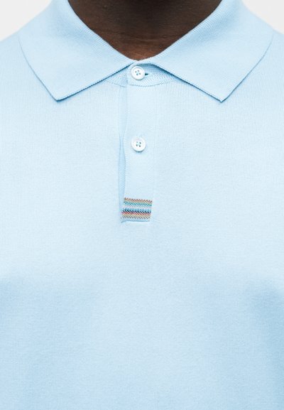 Gros plan sur une personne portant un polo bleu clair avec deux boutons blancs et un petit détail rayé multicolore sous les boutons.