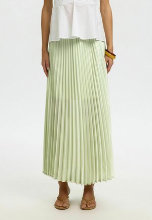 Kvinne iført en lys grønn plissert midiskjørt, hvit ermeløs topp, brune sandaler og to armbånd på høyre håndledd mot hvit bakgrunn.