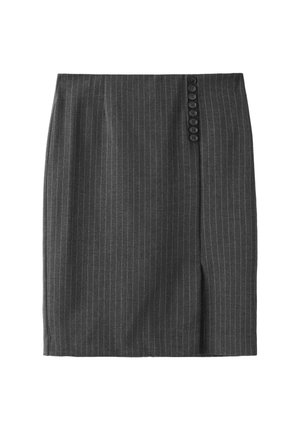 Femme debout en jupe grise à fines rayures, haut blanc à manches longues, talons noirs, tenant un sac à main noir, sur fond gris uni.