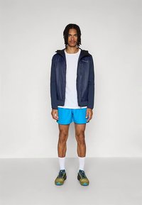 Vyras su pintomis plaukais, vilkintis tamsiai mėlyną švarką, baltą marškinėlį, ryškiai mėlynas šortus, baltas kojines ir žalias bėgimo batus ant balto fono.