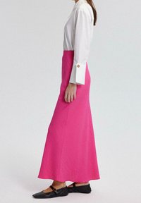 Maxirock in lebhaftem Pink mit strukturiertem Stoff; kombiniert mit einer taillierten weißen Bluse mit langen Ärmeln und goldenen Knopfdetails. Schwarze Ballerinas.