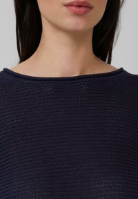 Das untere Gesicht und der Hals einer Frau mit glattem, dunklem Haar, die ein navyblaues, strukturiertes Strickoberteil mit rundem Ausschnitt trägt.