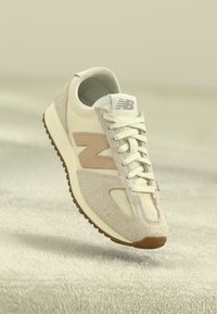 Beige Sportschuh mit einer strukturierten Oberseite, leichtem Mesh und einem auffälligen beige Logo. Verfügt über weiße Schnürsenkel und eine braune Gummisohle.