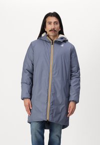 Cappotto impermeabile grigio con cappuccio, dotato di una cerniera gialla e bianca, polsini elasticizzati e una morbida fodera in pelliccia. Vestibilità casual fino al ginocchio.