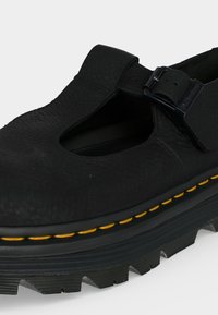 Chaussure Mary Jane en cuir noir avec une semelle épaisse nervurée, une couture jaune et une bride ajustable à boucle sur le dessus.