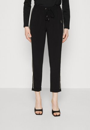 Femme porte un pantalon noir avec taille à cordon et bordure de strass sur les côtés, assorti à des talons ouverts noirs et un haut noir à manches longues.