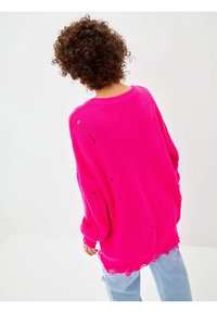 Personne aux cheveux bouclés portant un pull en maille rose vif usé et un jean bleu clair, de dos sur un fond uni.