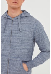 Sweatshirt à capuche bleu zippé en tissu doux et texturé. Il dispose d'une capuche, de cordons de serrage et de poches avant avec des poignets côtelés.