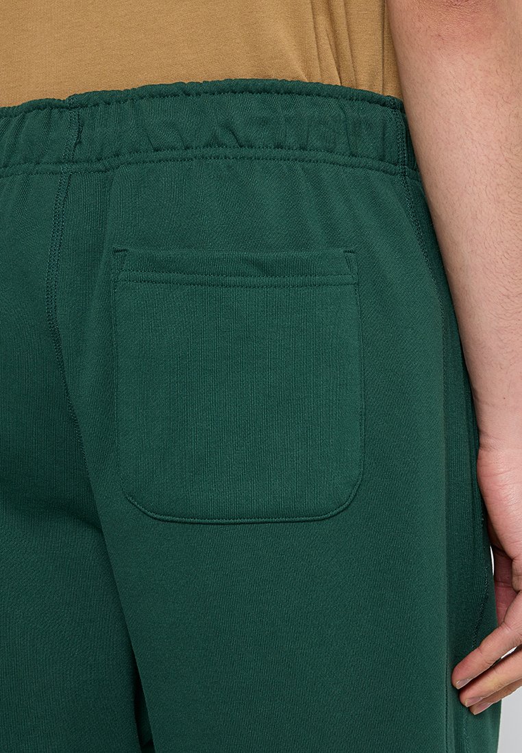 Ingrandimento di una persona che indossa pantaloni sportivi verde scuro con vita elastica e tasca posteriore, con la mano poggiata accanto alla gamba.