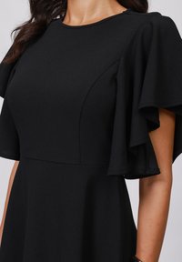 Robe noire avec des manches flottantes, un corsage ajusté et une texture de tissu lisse. Présente des coutures courbes et un col rond.